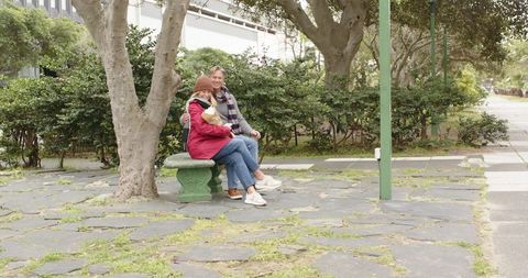 Senior couple sitting on green stone bench in urban park, woman holding grocery bag and smiling
