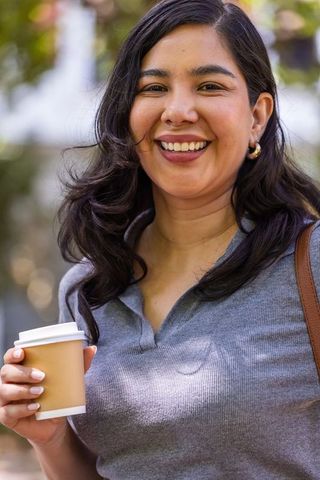 Smiling woman holding takeaway coffee outdoors wearing gray knit top and leather bag