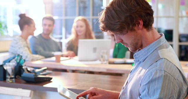 This image showcases a young professional focused on his tablet in a modern and vibrant office setting. In the background, team members are engaged in discussion, reflecting coworking and collaboration. It is perfect for content related to modern business, technology in the workplace, teamwork, collaborative environments, and digital tools.