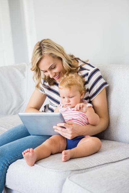 Young mother and baby girl sitting on couch using digital tablet together. Perfect for themes related to family bonding, modern parenting, technology in education, and home lifestyle. Ideal for use in advertisements, parenting blogs, educational content, and social media posts promoting family activities and digital learning.