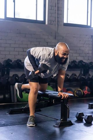 African american man performing one-arm dumbbell row on bench, smiling in industrial gym