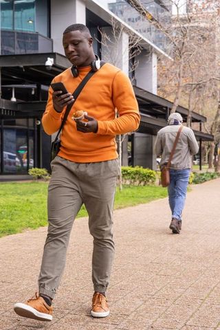 Mature african american man walking and checking phone while holding croissant in city park