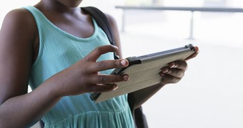 Girl using tablet in bright hallway wearing teal dress and backpack, hands tapping screen