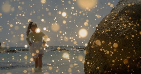 Wading child at rocky shoreline with golden bokeh glow at dusk, serene coastal moment