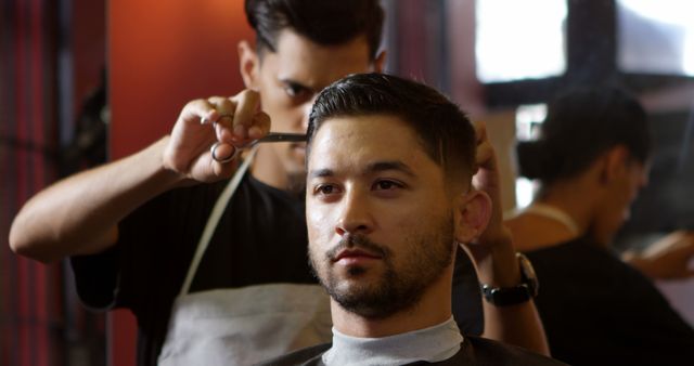 A professional barber of Middle Eastern ethnicity is giving a haircut to a young Asian man in a barbershop, with copy space. Precise grooming techniques are showcased as the barber attentively styles the client's hair.