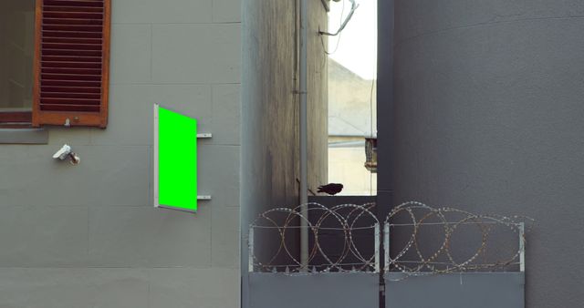 Perfect for showcasing urban safety and security themes, promotional advertisements in a city setting, or visual effects needing a blank green screen display. Image depicts a blank advertisement board affixed to a building in an alley, secured by a barbed wire fence with visible surveillance camera, embodying a gritty urban environment.