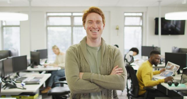 Man with red hair smiling confidently with arms crossed in a modern, bright office environment. Coworkers in the background, working at computer screens and collaborating. Ideal for use in marketing materials promoting positive workplace culture, modern office settings, teamwork, and professional services.
