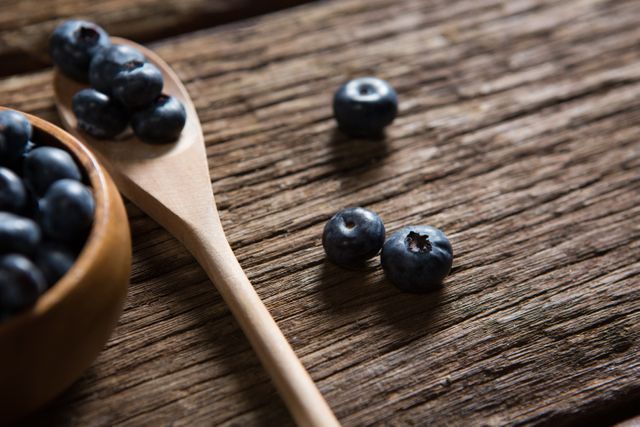 This image shows fresh blueberries on a rustic wooden table, with some in a wooden spoon and bowl. Ideal for use in food blogs, healthy eating articles, organic food promotions, and recipe websites.