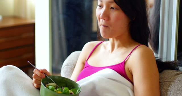 Woman is enjoying a healthy salad while relaxing on a couch indoors. She appears content and at ease, possibly taking a break. This image can be used for promoting healthy eating habits, wellness blogs, relaxation or leisure time content, lifestyle articles, and diet-related marketing materials.