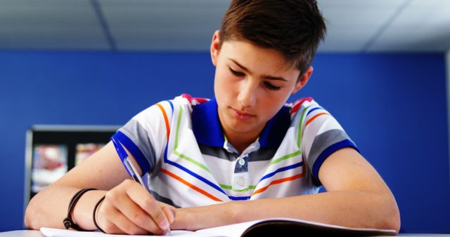 Teen boy concentrates on writing in notebook while seated at desk, wearing casual attire with striped shirt. Background features blue wall, typical classroom setting. Ideal for illustrating topics in education, study practices, school environment, youth focus, or academic achievement.