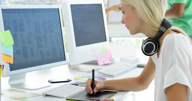 Young blonde designer using digital tablet at modern workspace. Wearing headphones, she is working on computer with creative tools surrounding her. Can be used to depict creative processes, freelancing, graphic design, modern technology, and professional work environments.