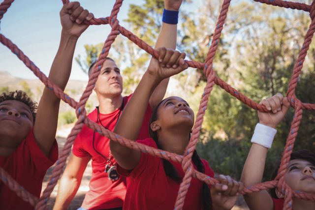 Trainer assisting kids climbing a net during an outdoor obstacle course in a boot camp. Ideal for use in articles about fitness, teamwork, summer camps, children's physical activities, and outdoor adventures.