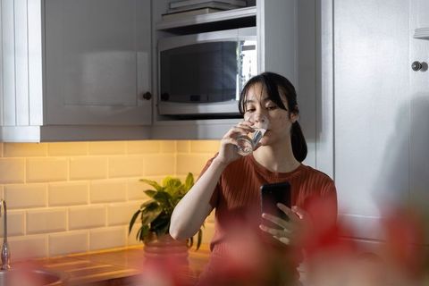 Asian woman sipping water and checking smartphone in cozy modern kitchen
