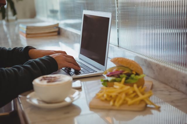 Cropped hands of man using laptop by coffee cup and French fries at counter in cafe