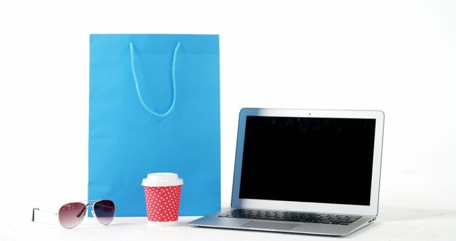 A blue shopping bag, a red cup with a lid, sunglasses, and an open laptop are arranged on a white surface, with copy space. This setup suggests a blend of leisure and work, representing online shopping or a casual business environment.