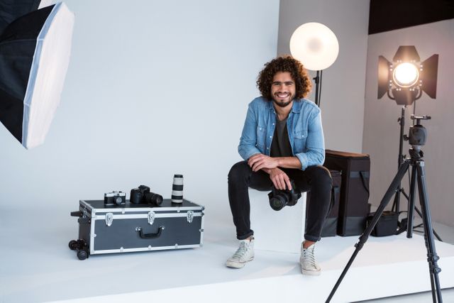 Photographer sitting on stool in studio surrounded by camera equipment and lighting. Ideal for use in articles about photography, creative professions, studio setups, or professional portraits.