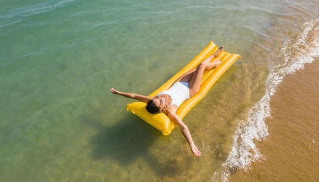 Sunbathing woman floating on yellow air mattress at clear turquoise shoreline, aerial view