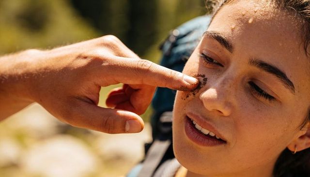 Teen hiker laughing while friend smearing mud on cheek during sunny trail break