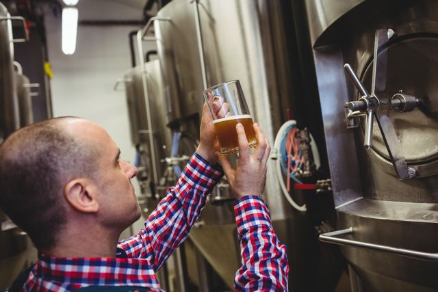 Brewery worker inspecting beer quality in glass, ensuring standards in industrial brewing process. Ideal for use in articles about craft beer production, quality control in beverage industry, professional brewing techniques, and industrial manufacturing processes.
