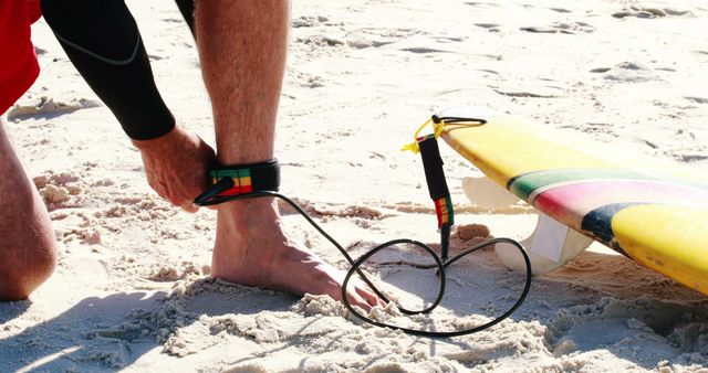 A surfer, a young or middle-aged Caucasian male, is attaching a leash to his ankle before heading out to surf, with copy space. His surfboard is brightly colored, and the sandy beach setting suggests a day of recreational surfing ahead.