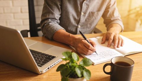 Man writing notes at home office desk with laptop, notebook, coffee mug and plant