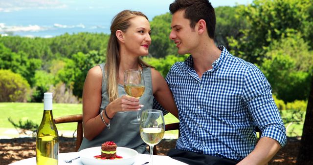 A young Caucasian couple enjoys a romantic outdoor meal with wine, with copy space. Their intimate moment and scenic backdrop suggest a special occasion or celebration.