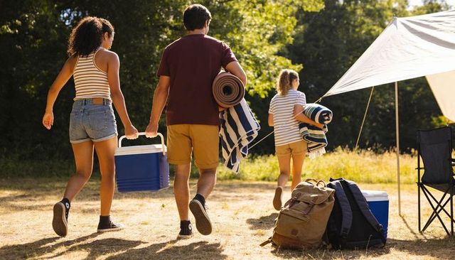 Friends walking carrying cooler and rolled mats toward tarped campsite in sunlit meadow