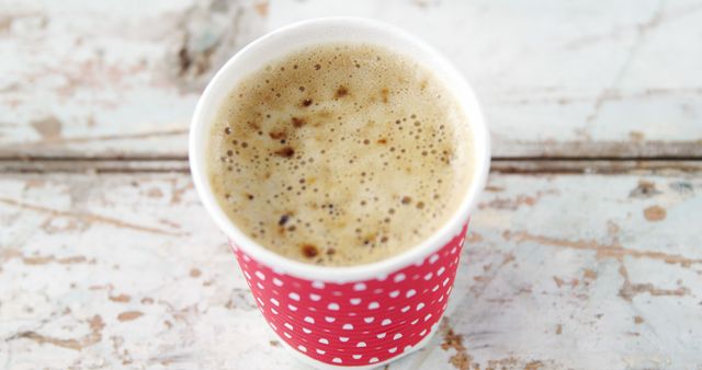A cup of frothy coffee in a red polka-dot paper cup sits on a rustic surface, with copy space. Its inviting appearance suggests a warm, comforting beverage option for coffee enthusiasts.