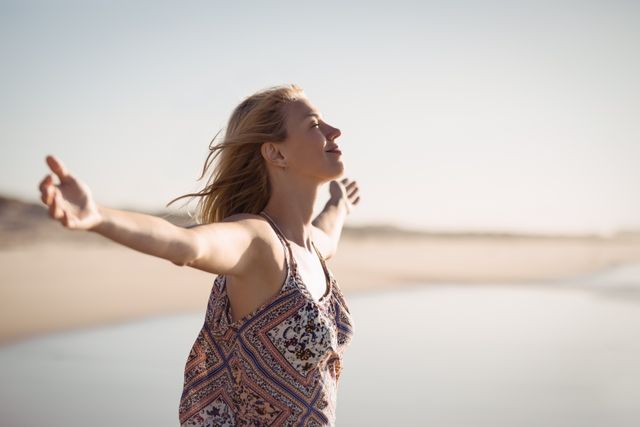 Woman enjoying a sunny day at the beach with arms outstretched, symbolizing freedom and happiness. Ideal for use in travel brochures, wellness blogs, lifestyle magazines, and advertisements promoting relaxation and outdoor activities.