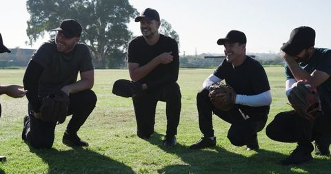 Men kneeling and laughing on baseball field holding gloves, showing team camaraderie