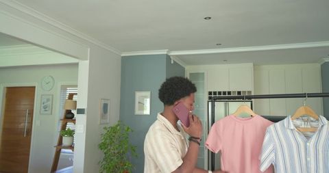African american man choosing shirt while talking on smartphone at clothing rack in home
