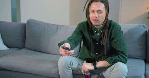 African american man sitting on sofa holding remote and drink bottle wearing smartwatch