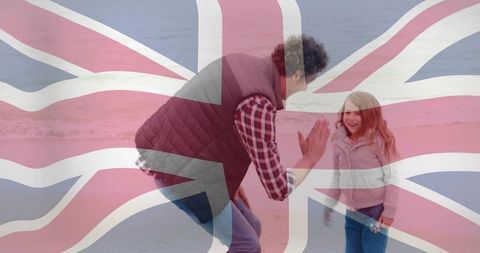 Father high-fiving daughter on beach with translucent union jack overlay, family seaside moment