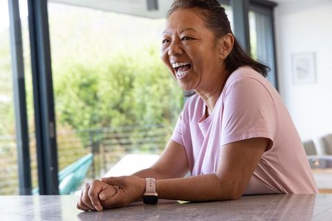 Senior asian woman laughing at kitchen counter wearing smartwatch and pink tee in bright home