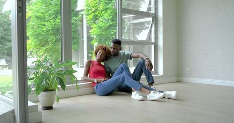 Diverse couple sitting on sunlit floor by large windows in minimalist modern home