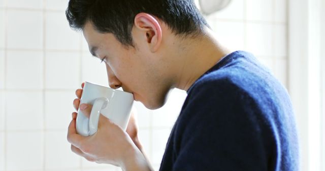 Young man sipping beverage from a white mug indoors. Picnicking subtle and relaxed mood making it suitable for themes related to relaxation, casual refreshment, coffee breaks, comfort, and daily routines. Useful for illustrating casual lifestyle, advertisement for drinks, or articles on relaxation techniques and taking breaks.