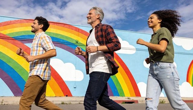 Three friends jogging along vibrant rainbow mural on sunny urban street, casual attire
