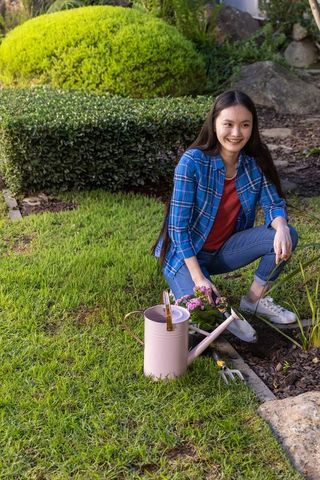 Smiling asian woman kneeling in sunny backyard gardening with pink watering can and flowers