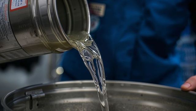 Pouring clear viscous liquid from metal can into stainless vat by technician in lab