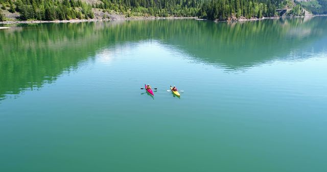 Two kayakers paddle on a tranquil mountain lake surrounded by lush greenery. The clear, calm water reflects the natural beauty of the towering mountains and dense forest. Ideal for promoting outdoor adventure, nature exploration, and water sports. Perfect for use in travel brochures, tourism promotions, and advertisements for outdoor gear.