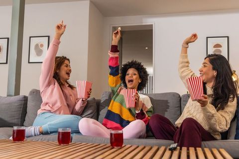 Diverse friends cheering and sharing popcorn on gray sofa during cozy movie night