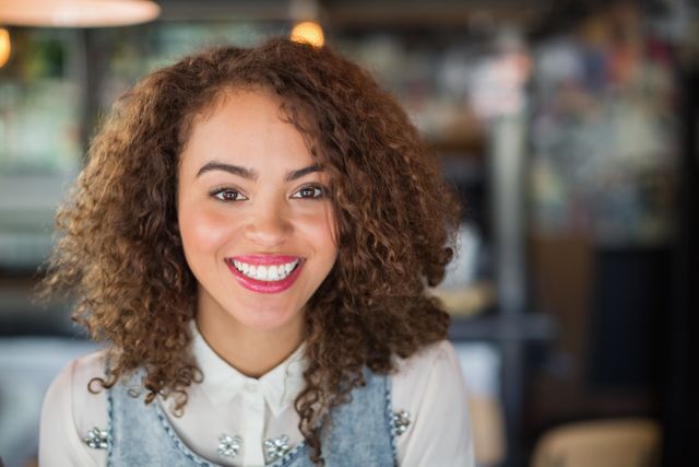 This image features a young woman with curly hair smiling warmly in a casual pub environment. Ideal for use in lifestyle blogs, social media posts, advertisements for pubs or restaurants, and articles about socializing and leisure activities.