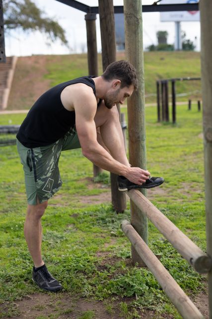 Side view of a Caucasian man wearing a black vest and camo shorts with his foot up on a fence tying his shoe at an outdoor gym during a bootcamp training session, with outddor gym equipment in the background