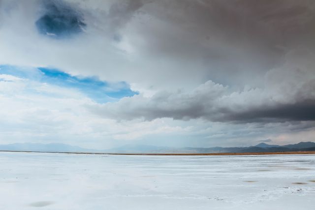 A landscape featuring a wide, expansive salt flat under a stormy sky, with distant mountains visible on the horizon. Ideal for use in travel blogs, nature documentaries, and outdoor adventure content to highlight serene and untouched scenic beauty, vast natural wastelands, and arid environments.