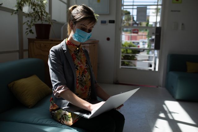 Caucasian woman sitting on a couch in an office, wearing a facemask and typing on her laptop. Ideal for use in articles about remote work, health and safety in the workplace, pandemic precautions, and professional settings.