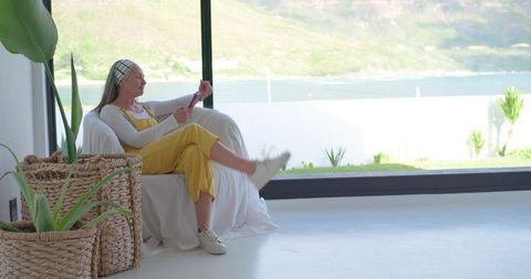 Mature woman relaxing in sunlit modern living room wearing yellow overalls using smartphone