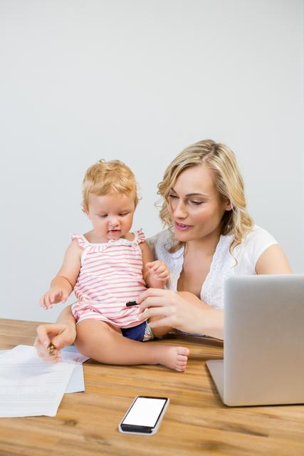 Mother multitasking with baby girl and laptop at home, ideal for illustrating work-life balance, remote work, parenting, and family life. Useful for articles, blogs, and advertisements related to motherhood, working from home, and child care.