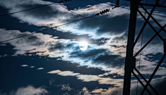 Sun breaking through layered clouds over power transmission tower with silhouetted lines