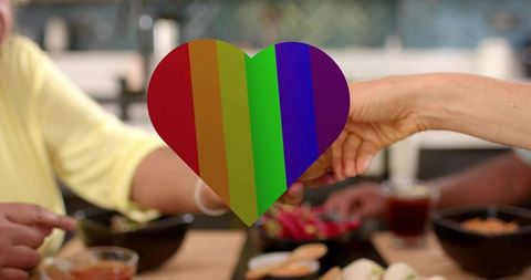 Senior friends sharing brunch, passing buns, rainbow heart symbolizing pride and inclusion
