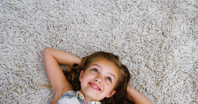 A young Caucasian girl lies on a fluffy carpet, looking up with a dreamy expression, with copy space. Her relaxed pose and happy demeanor suggest a moment of leisure or daydreaming at home.